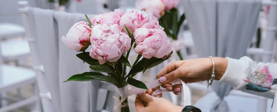 Handen die roze pioenrozen aan een bruiloftstoel binden als onderdeel van de decoratie voor de ceremoniezitplaatsen.